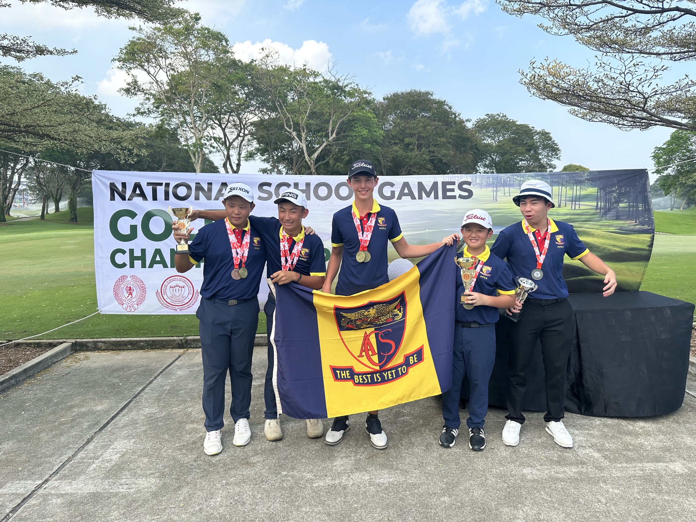 Five young golfers with medals and trophies hold a school flag in front of a National School Games Golf banner.